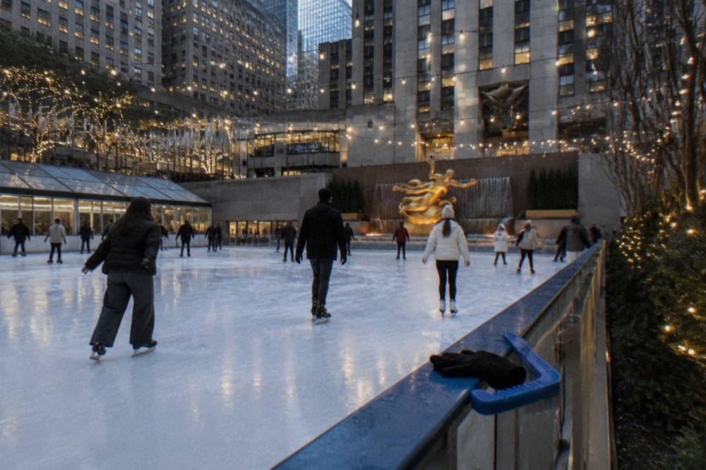 Ice skaters gliding on an outdoor rink surrounded by holiday lights and city towers in early evening