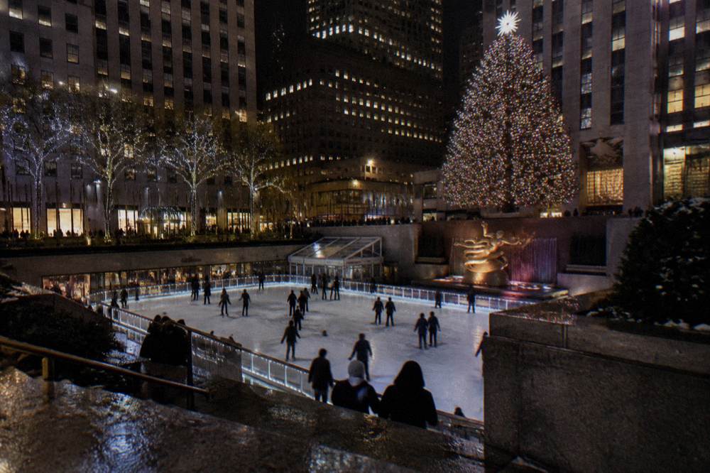 An illuminated ice rink beneath a tall decorated evergreen in a plaza at night