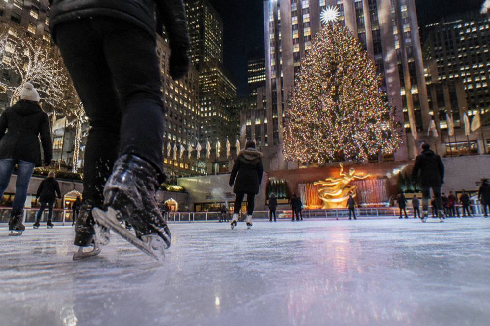 A tall lit Christmas tree rising above an ice rink in a stone plaza at night