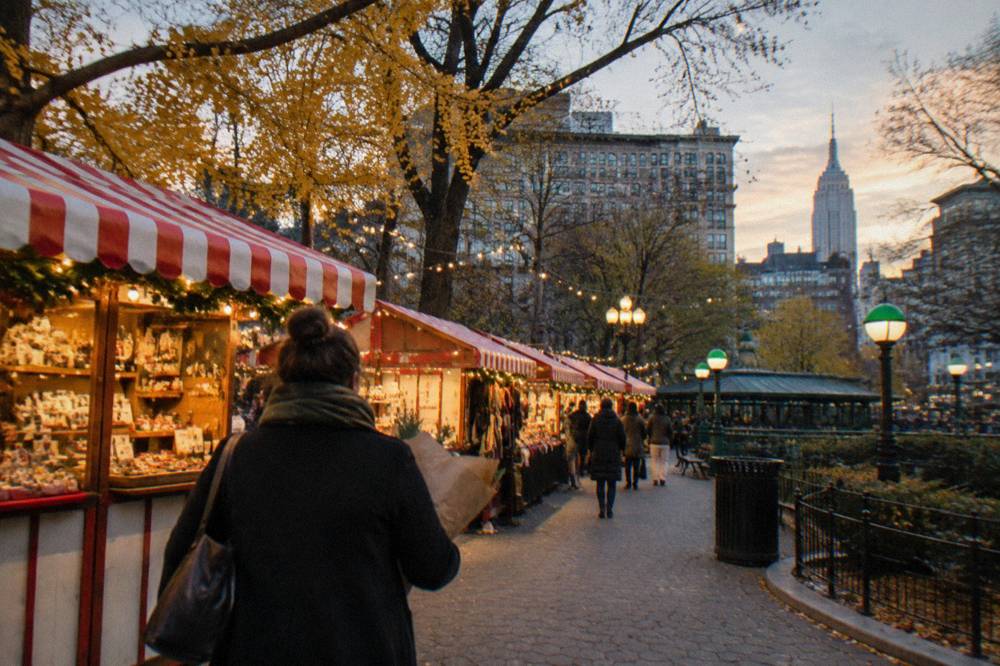 Holiday market kiosks with warm string lights lining a park path in the early evening