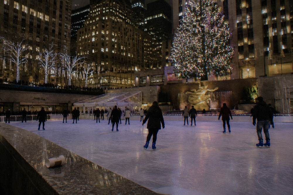 Skaters circling an illuminated outdoor rink surrounded by city towers after dark