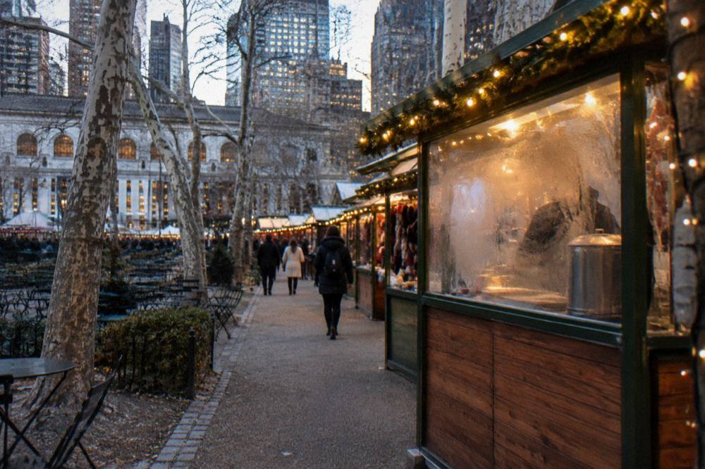 Holiday market kiosks with string lights along the edge of Bryant Park in the early evening
