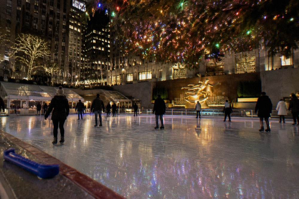 An ice rink beneath a glowing Christmas tree and surrounding city towers at night