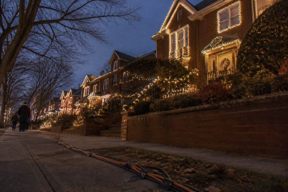 A residential Brooklyn block at night with holiday light displays glowing along both sidewalks
