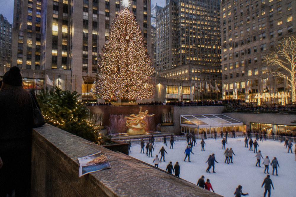 A giant lit Christmas tree above a plaza ice rink in Midtown Manhattan at night