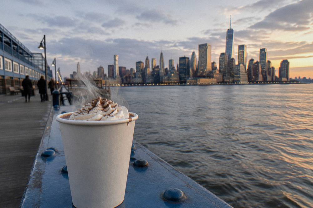 A cup of hot cocoa topped with whipped cream resting on a yacht railing with the Manhattan skyline behind it
