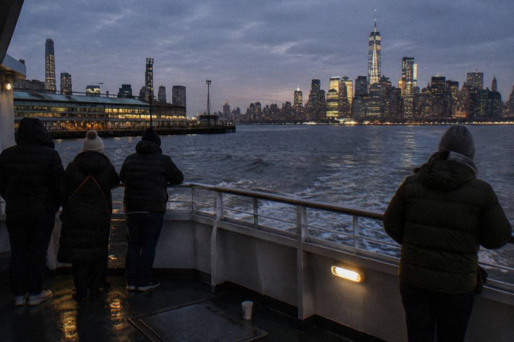 Passengers wrapped in winter coats looking out from a yacht deck toward the lit Manhattan skyline