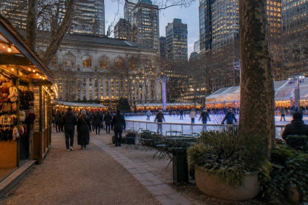 An ice rink surrounded by wooden market kiosks and strung lights beside a park boundary of tall buildings