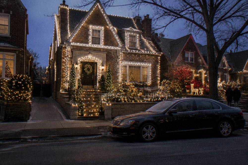 A dark private car waiting at a Brooklyn curb beneath rows of illuminated holiday displays