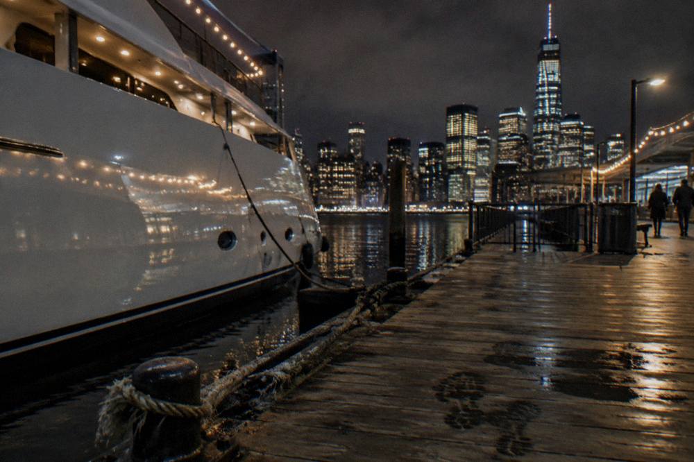 A yacht strung with white holiday lights moored at a Hudson River pier with the Manhattan skyline glowing behind it