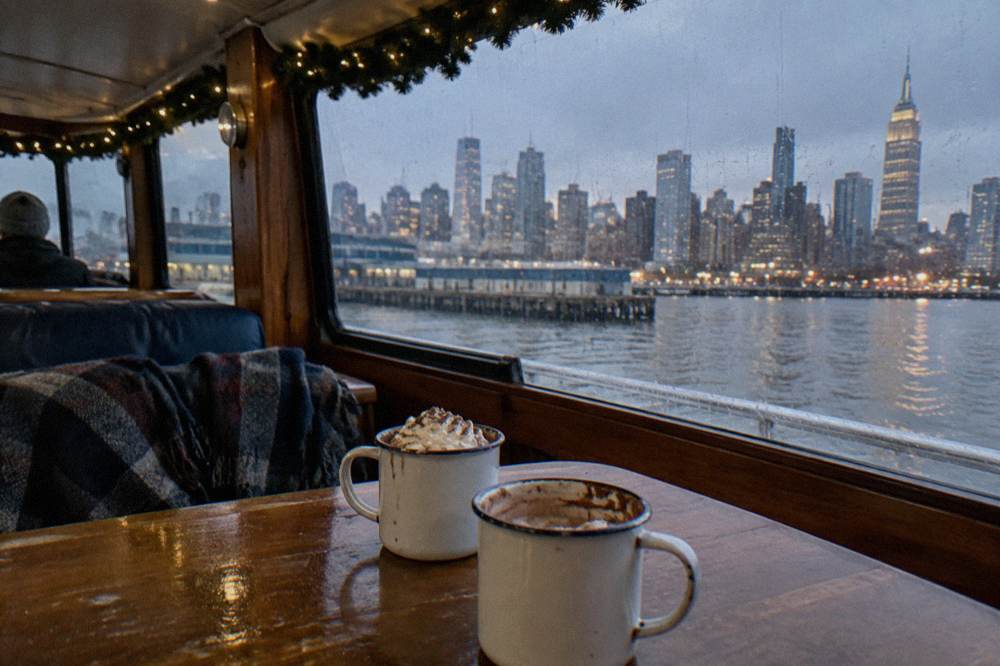 Two mugs of hot cocoa topped with whipped cream on a wooden table with blurred city lights through a window