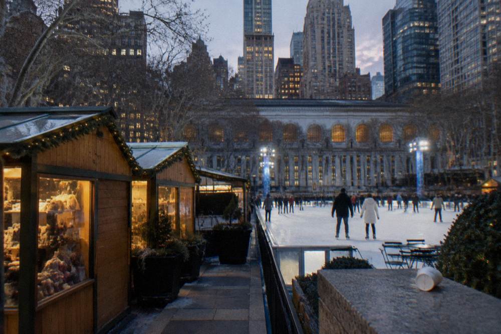 An ice rink framed by wooden market stalls and Midtown skyscrapers at dusk