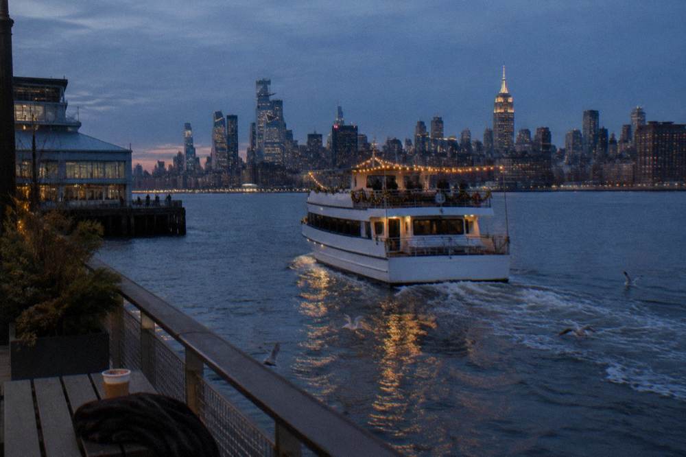 A decorated yacht with warm holiday lights along its railings cruising past the Manhattan skyline at dusk