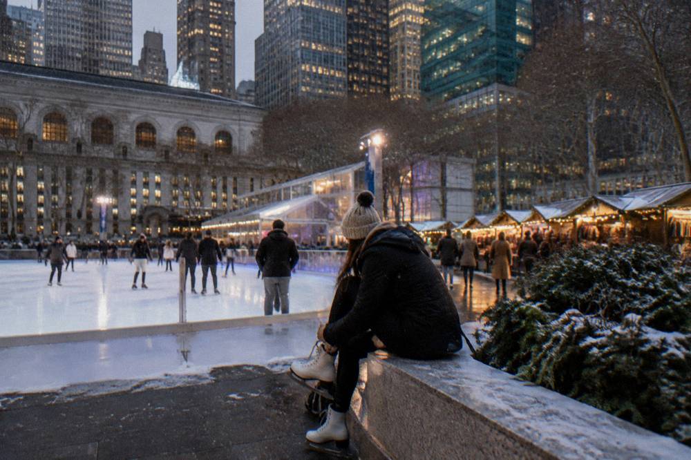 An ice rink surrounded by tall buildings and strings of lights beside festive wooden market stalls in a Manhattan park