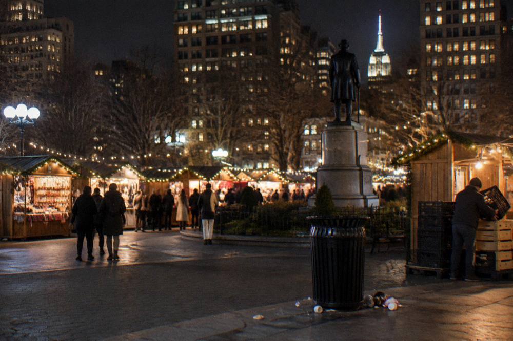 Festive wooden holiday market stalls with strings of lights glowing along the edge of a Manhattan park at night