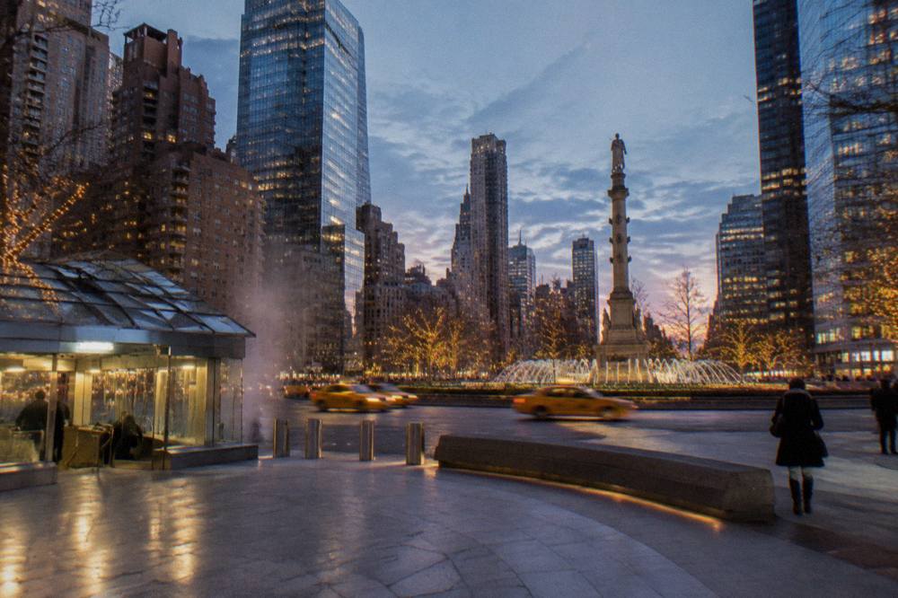 Wide view of Columbus Circle at dusk with holiday lights on buildings and a subway station entrance