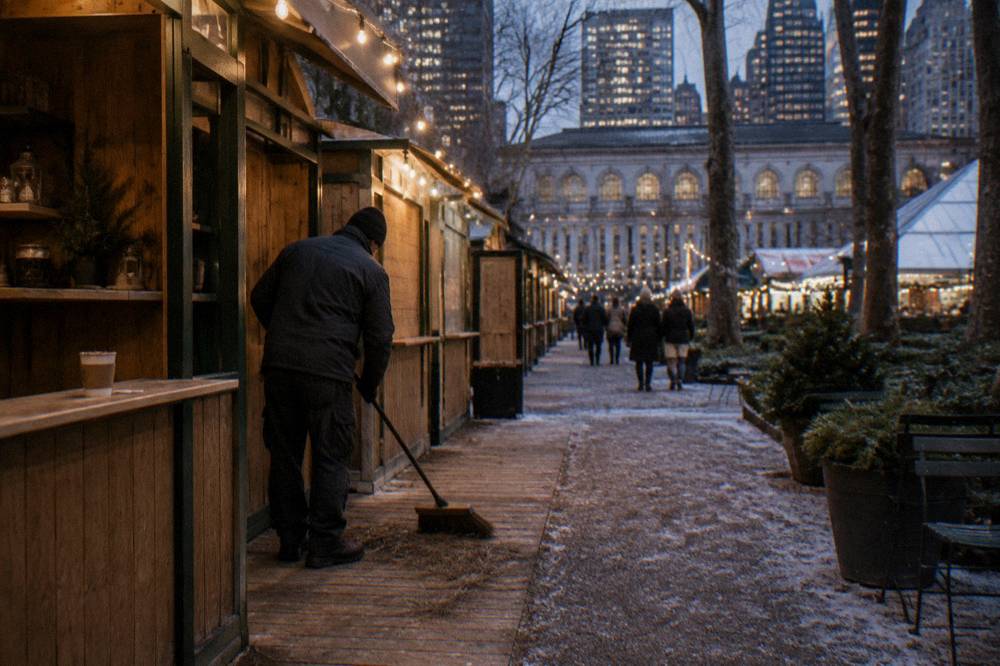 Row of wooden holiday market kiosks with glowing string lights in a New York park after dark