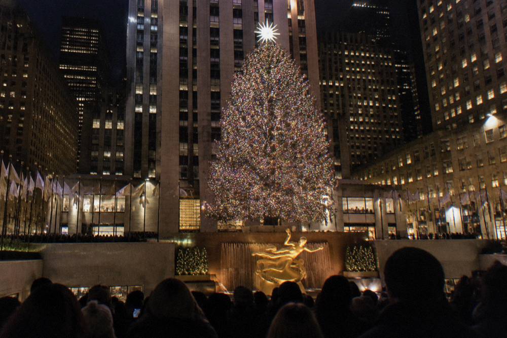 A giant lit Christmas tree glowing above the plaza surrounded by golden angel sculptures and flags