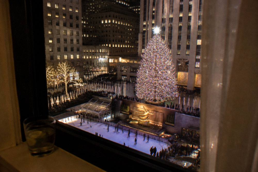 A festive hotel window overlooking a plaza with a lit Christmas tree at night