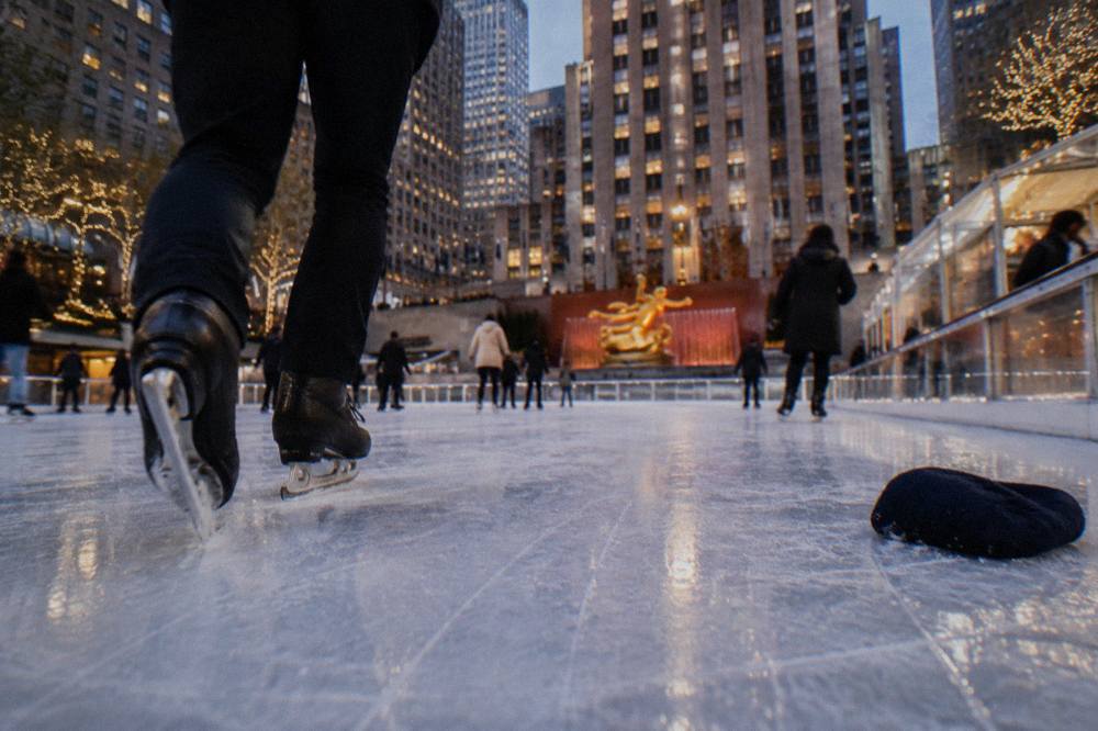 Close view of an outdoor ice rink surface with skaters circling beneath glowing tower buildings at dusk