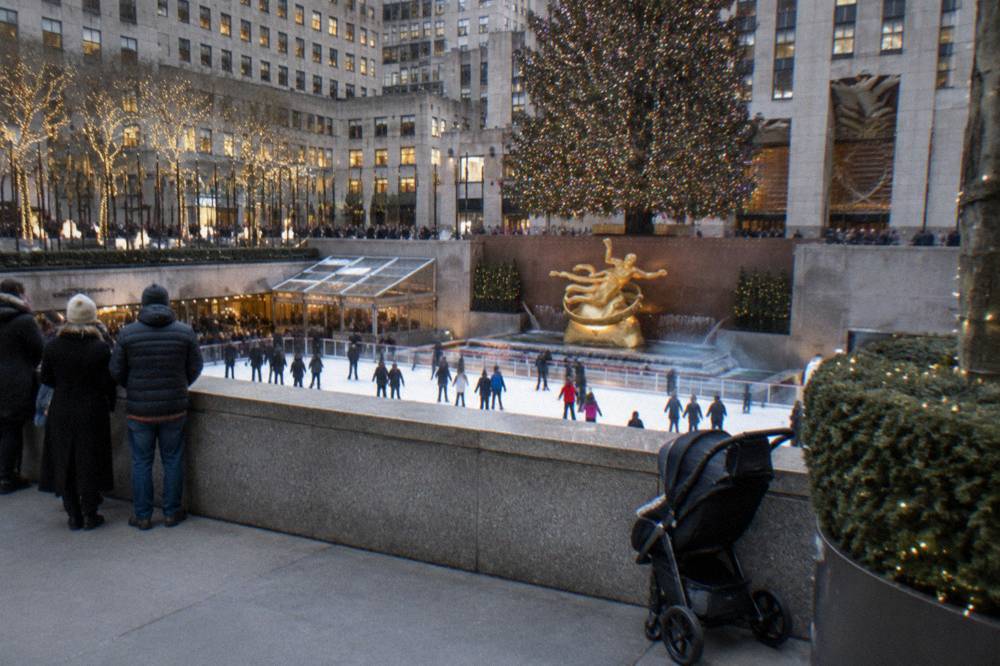 Wide view of a plaza rink surrounded by winter shoppers and stone buildings with a large decorated tree in the background