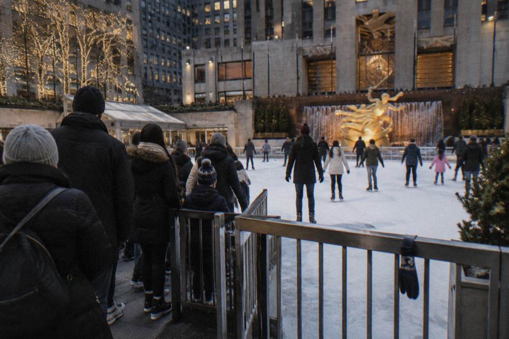 Skaters of mixed ages circling a small lit rink in the evening with a queue forming at the gate beside the ice