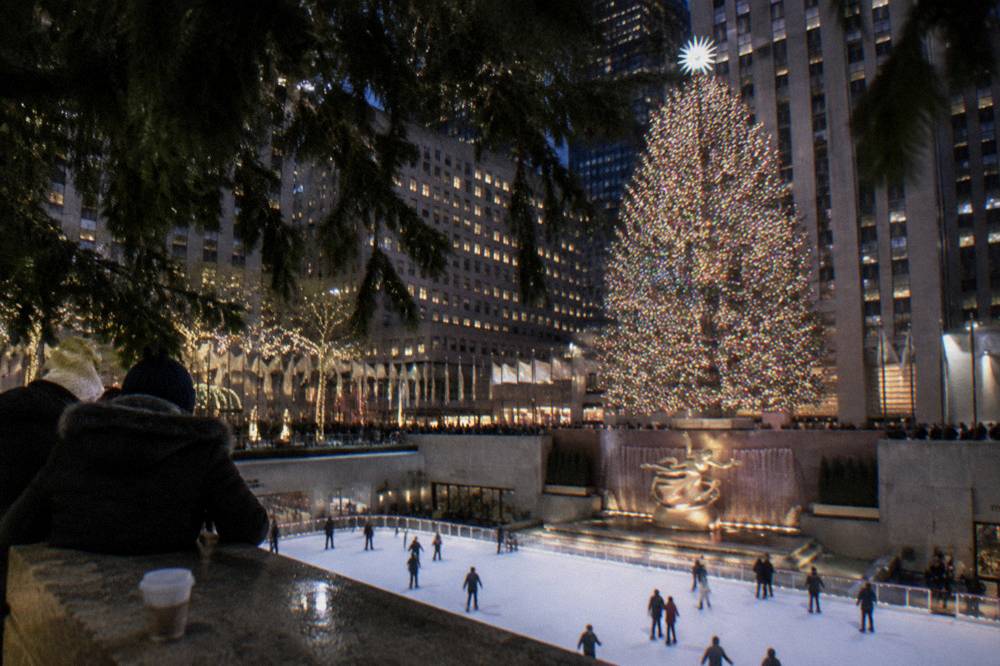 A giant lit Christmas tree towering over a plaza rink with skaters below and spectators leaning on the railings