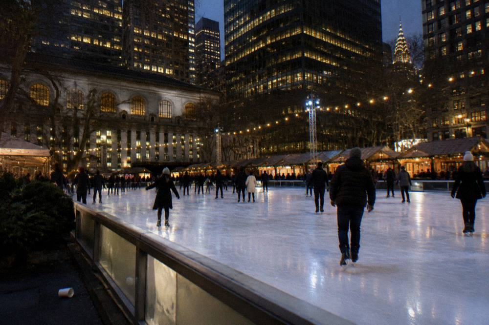 An outdoor rink ringed by wooden market huts and tall buildings lit for the holidays at night