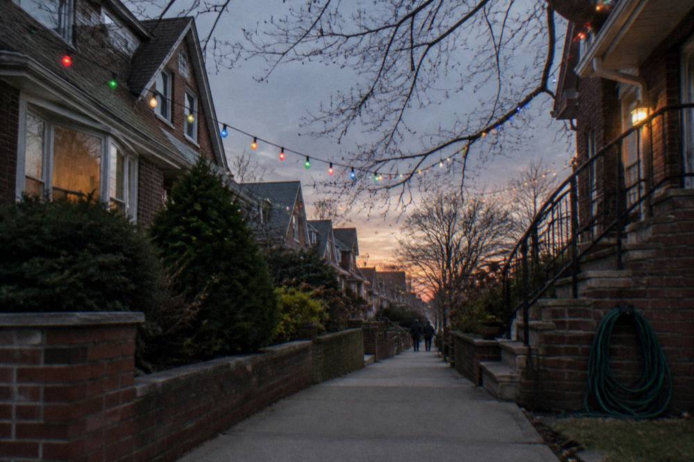 Residential Brooklyn street at dusk with rows of Christmas lights glowing above the sidewalk